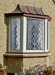 A house window with diamond-patterned leaded glass, set in a light-colored textured wall, surrounded by flowers below.